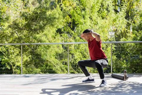 Man performing squats hands-behind-head on deck by railing, red shirt, mat and Stock Photos