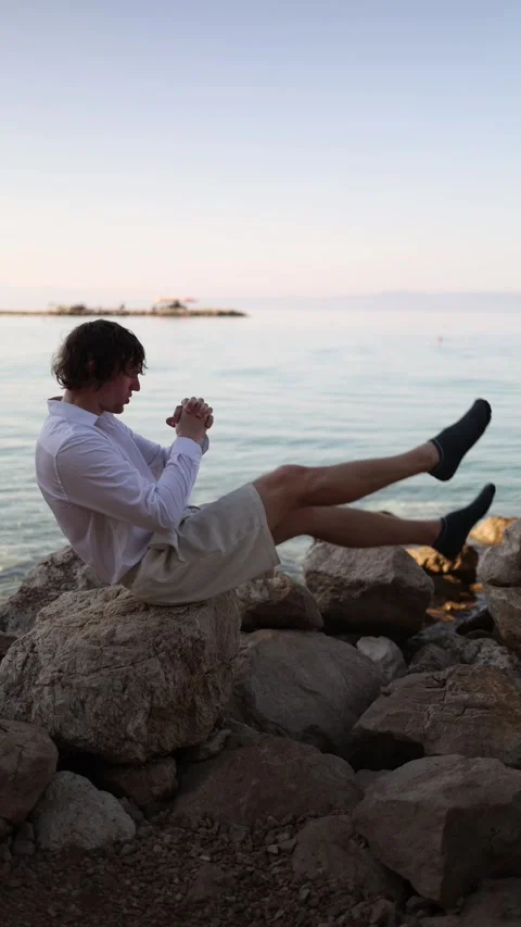 Man performs seated leg raises on coastal rocks during outdoor core workout Stock Footage 341621856