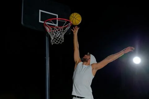 A man performs a slam dunk on a basketball court at night under the glare o.. Stock Photos