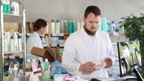 Man pharmacist uses computer at workplace. Girl assistant in background Stock Footage 317994225