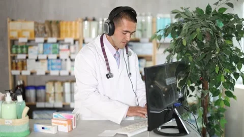Man pharmacist works at computer using headset, girl assistant in background Stock Footage 332735825