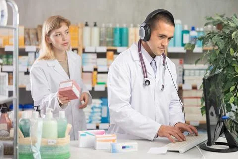 Man pharmacist works at computer using headset, girl assistant in background Stockfoto's