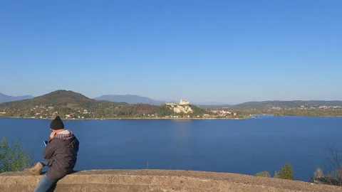 Man with phone sitting on wall overlooking lake maggiore and fortress Stock-Footage 280882526