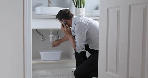 Man Phoning Emergency Plumber After Water Leak At Bathroom, Home Stock Footage 120893823