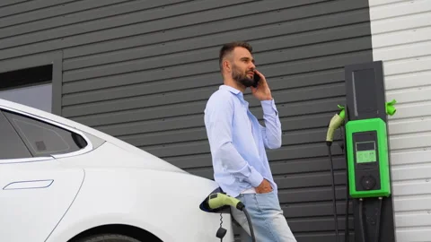 A man phoning while charging car at electric vehicle charging station Stock Footage 251765376
