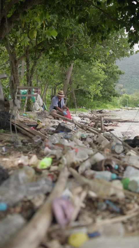 Man picking up plastic waste from a polluted tropical beach beneath trees 動画素材 343573975