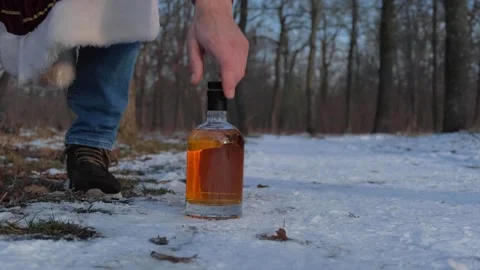 A man picks up a bottle of whiskey standing in the snow in a forest Stock Footage 318357171