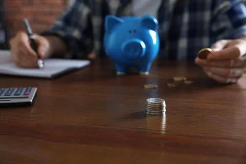 Man with piggy bank counting money at wooden table, focus on stack of coins Stock Photos
