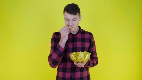 Man in a pink plaid shirt eats potato chips with glass bowl on yellow background Stock Footage 132381318