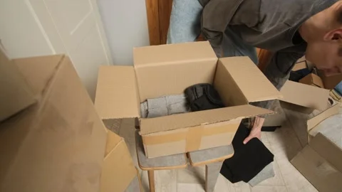 A man places folded clothes into an open cardboard box while preparing for a Stock Footage 303202025