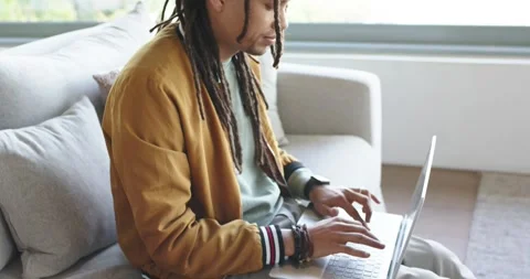 Man is placing laptop in lap on gray sofa near window, is typing for remote work Vídeos de archivo 341240744