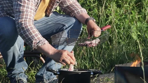 Man placing raw meat on grill for barbecue, outdoor cooking by river Stock Footage 332307631