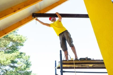 Man on platform extends to secure upper gable timber, yellow facade behind,.. Stock Photos