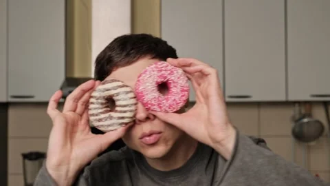 Man playfully holds two donuts over his eyes like glasses in a kitchen, making a Stock Footage 305736314
