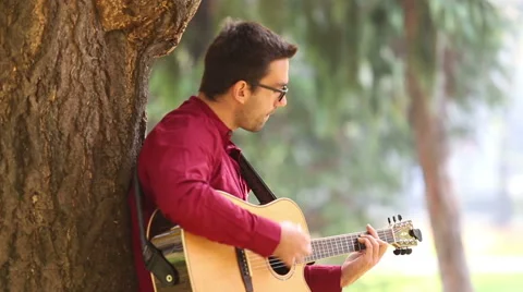 Man playing acoustic guitar leaning against a tree Stock Footage 59556515
