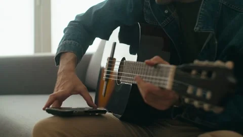 Man Playing Classical Guitar On Sofa While Using Smartphone App Stock Footage 336687217