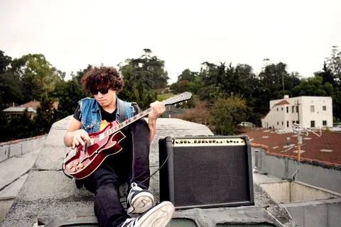 Man playing electric guitar while sitting on building against sky Fotos de archivo