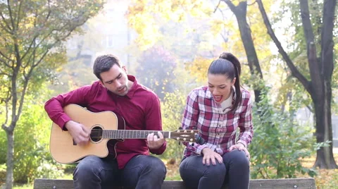 Man playing guitar and singing with women while sitting on bench in park Stock Footage 59556807