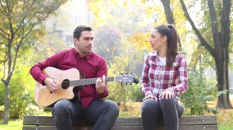 Man playing guitar and singing with women while sitting on bench in park Stock Footage 59556838
