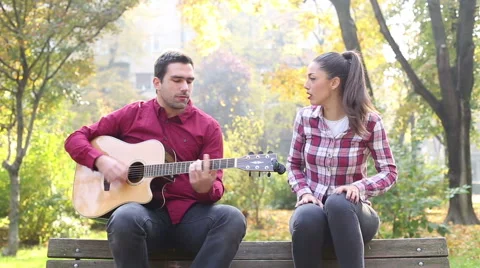 Man playing guitar and singing with women while sitting on bench in park Stock Footage 59556839