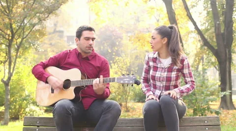 Man playing guitar and singing with women while sitting on bench in park Stock Footage 59556841