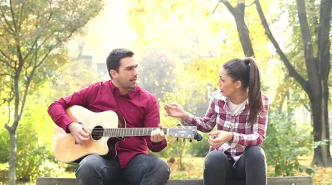 Man playing guitar and singing with women while sitting on bench in park Stock Footage 59556846