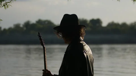 Man playing guitar by the river in serene woods during golden hour Stock Footage 319948036