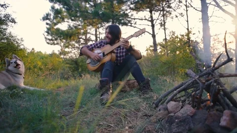 Man playing guitar sitting near the campfire in forest during sunset Vídeos de archivo 84422962