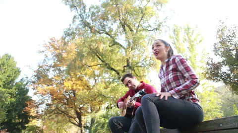 Man playing guitar while woman singing, sitting next to him on bench in park Stock Footage 59557036
