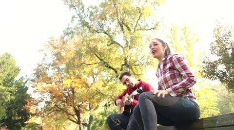 Man playing guitar while woman singing, sitting next to him on bench in park Stock Footage 59557037