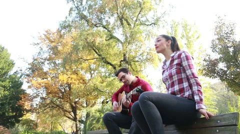 Man playing guitar while woman singing, sitting next to him on bench in park Stock Footage 59557038
