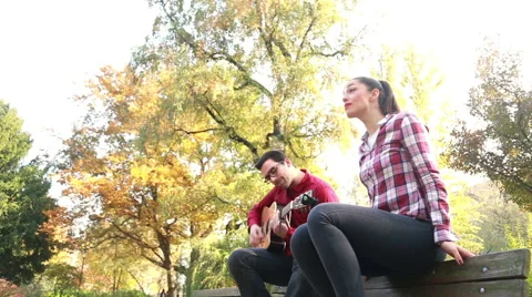 Man playing guitar while woman singing, sitting next to him on bench in park Stock Footage 59557043