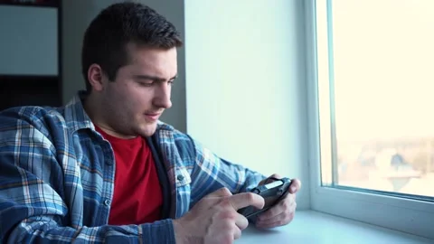A man playing a small console sitting by the window 스톡 동영상 167180414