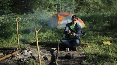 A man playing on a tablet in the game near the campfire and tents. In the forest Stock Footage 64312989