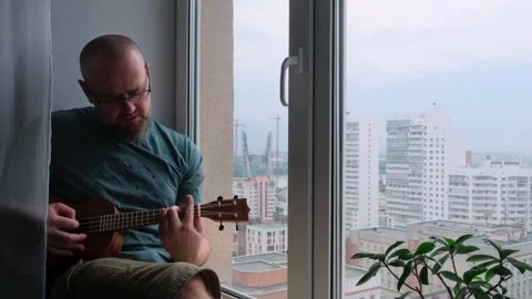 Man playing the ukulele sitting on the windowsill in a city apartment. Stock Footage 247604324