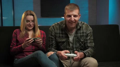 Man playing video games at concole while his girl play game on smartphone in Stock Footage 225653520