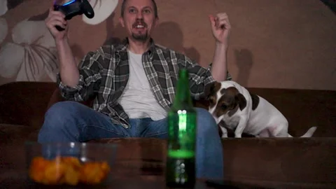 Man playing video games on sofa with dog and snacks at home. A man sitting .. Stock Footage 332416151