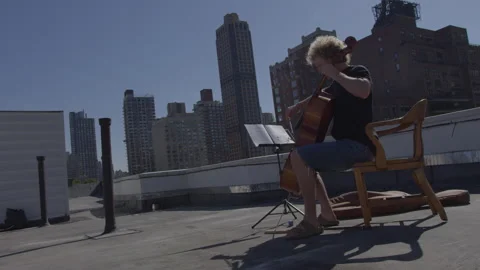 Man plays cello on rooftop while reading sheet music in urban setting Stock-Footage 312632381