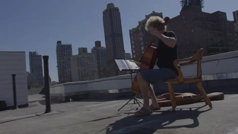 Man plays cello on rooftop while reading sheet music in urban setting Stock Footage 312639879