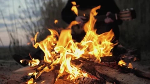 A man plays the guitar. In the foreground is a fire. Summer evening. Slow motion Stock Footage 130730539
