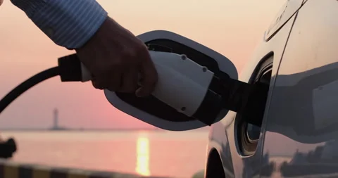 Man plugs charger into electric car socket against sunset Stock Footage