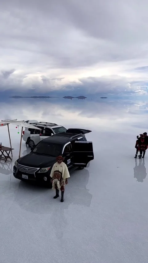 Man in Poncho Standing by SUV on Flooded Salar de Uyuni Salt Flat in Bolivia Видео 345071924