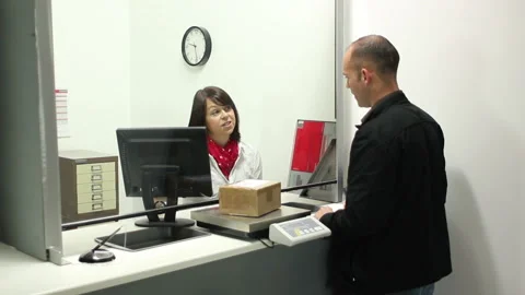 Man posting out a Parcel at the Post Office Counter - Female assistant serves Stock Footage 131841320