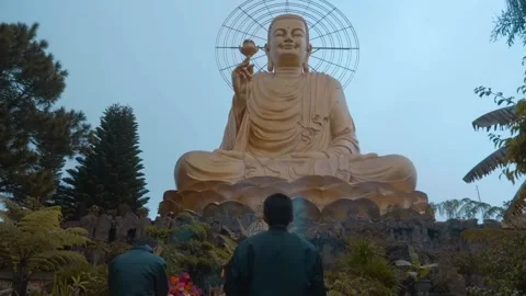 A man is praying in front of a massive golden Buddha statue with a neon halo Stock Footage 333339275