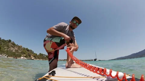 Man preparing for paddleboarding by securing leash to ankle on a board, in Stock Footage 313802272