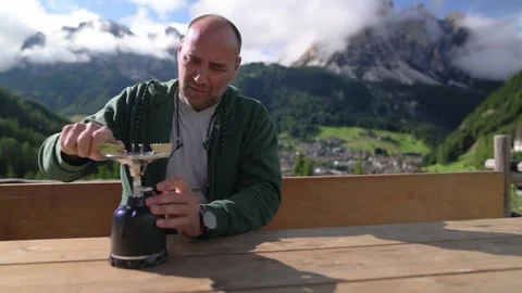 Man preparing tea with gas stove on wooden terrace with Dolomites village and 스톡 동영상 319412499