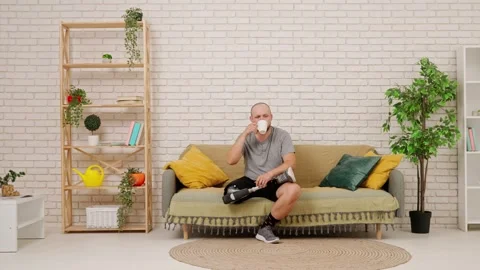 A man with a prosthetic leg is sitting on a sofa in a cozy living room, drinking Stock Footage 301072299