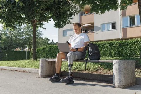 Man with a prosthetic leg using a laptop and smartphone while sitting outdoors Stock Photos