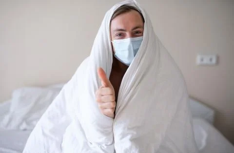 Man with a protective mask on his face wrapped in a blanket wakes up at home on Stock Photos