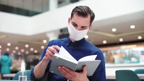 Man in a protective mask reads a book while sitting at a table in a cafe. Stock Footage 221126613